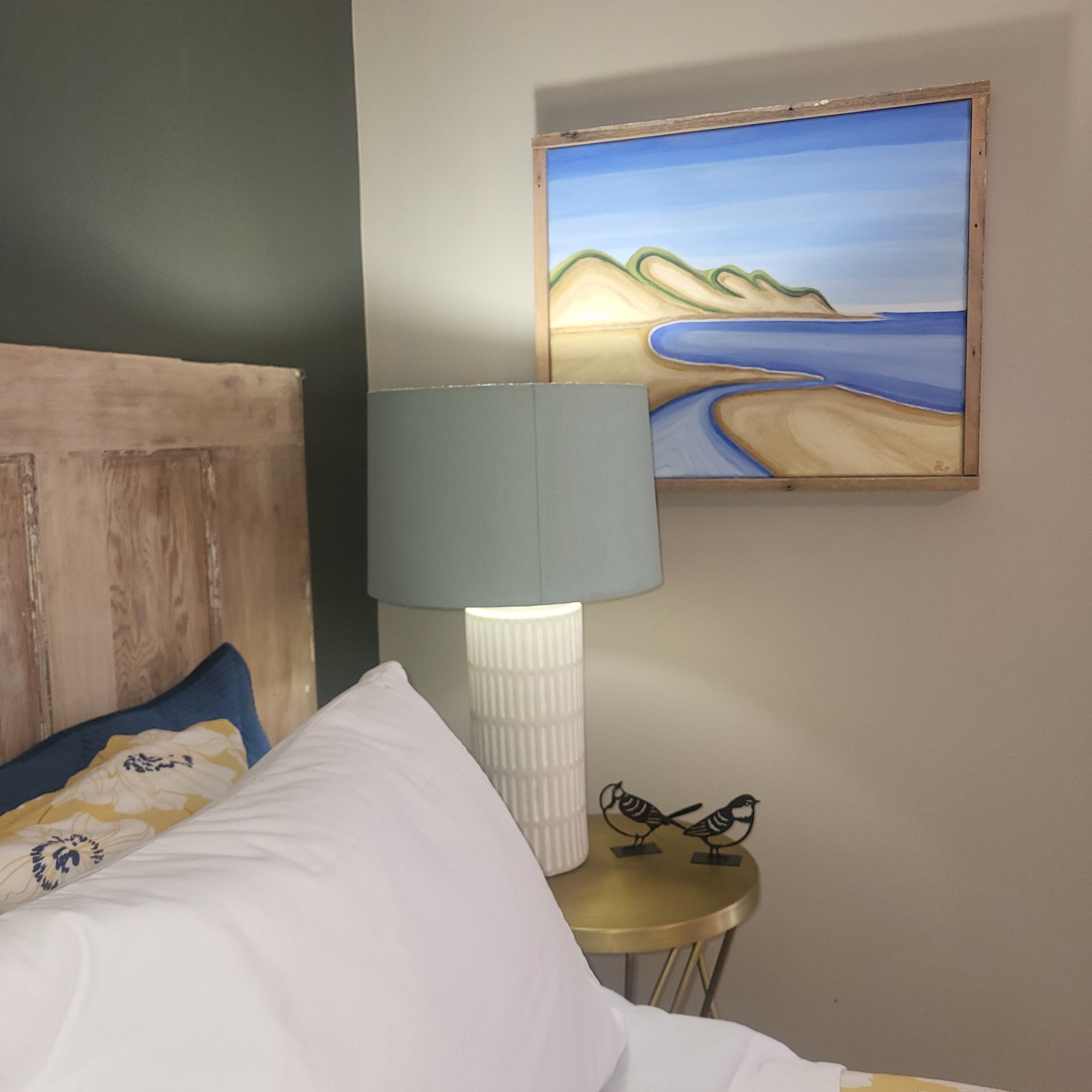 A bedroom with a bed, a lamp, a painting, and a small table with a bird sculpture.