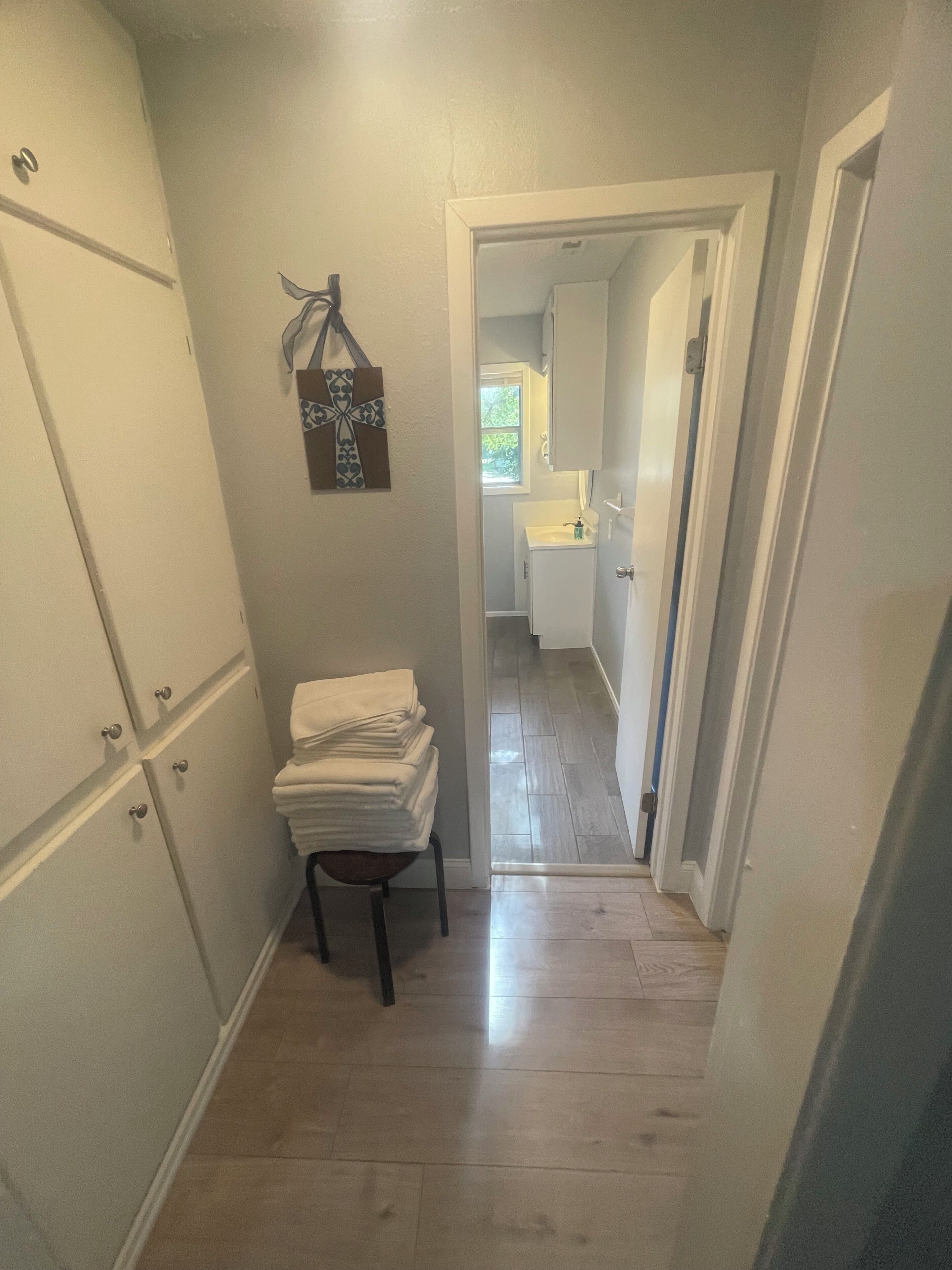 A narrow hallway with a wooden floor, a stack of white towels on a stool, and a door leading to a bathroom with a sink and window.