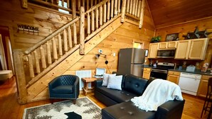Interior acolhedor de uma cabana com escada, sala de estar com sofá e poltrona, e cozinha. Uma placa de boas-vindas.