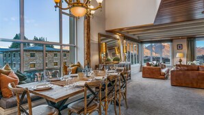 A spacious dining area with a high table set for six, surrounded by wooden chairs. A large mirror reflects the room, and a chandelier hangs above. The room has large windows offering a view of mountains and buildings. - The Ballarat Apartment (Queenstown)