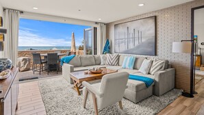 Una sala moderna con un sofá modular grande, una mesa de centro y un comedor con sillas. Hay un ventanal que ofrece vistas al océano.