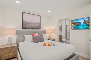 Une chambre à coucher bien rangée, avec un grand lit, deux tables de chevet, une télévision et une salle de bain visible à travers une porte ouverte.