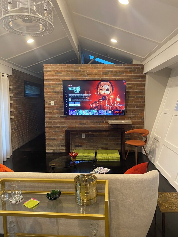A modern living room with a large flat-screen TV mounted on a brick wall, a glass coffee table with decorative items, a comfortable sofa, and a fireplace.