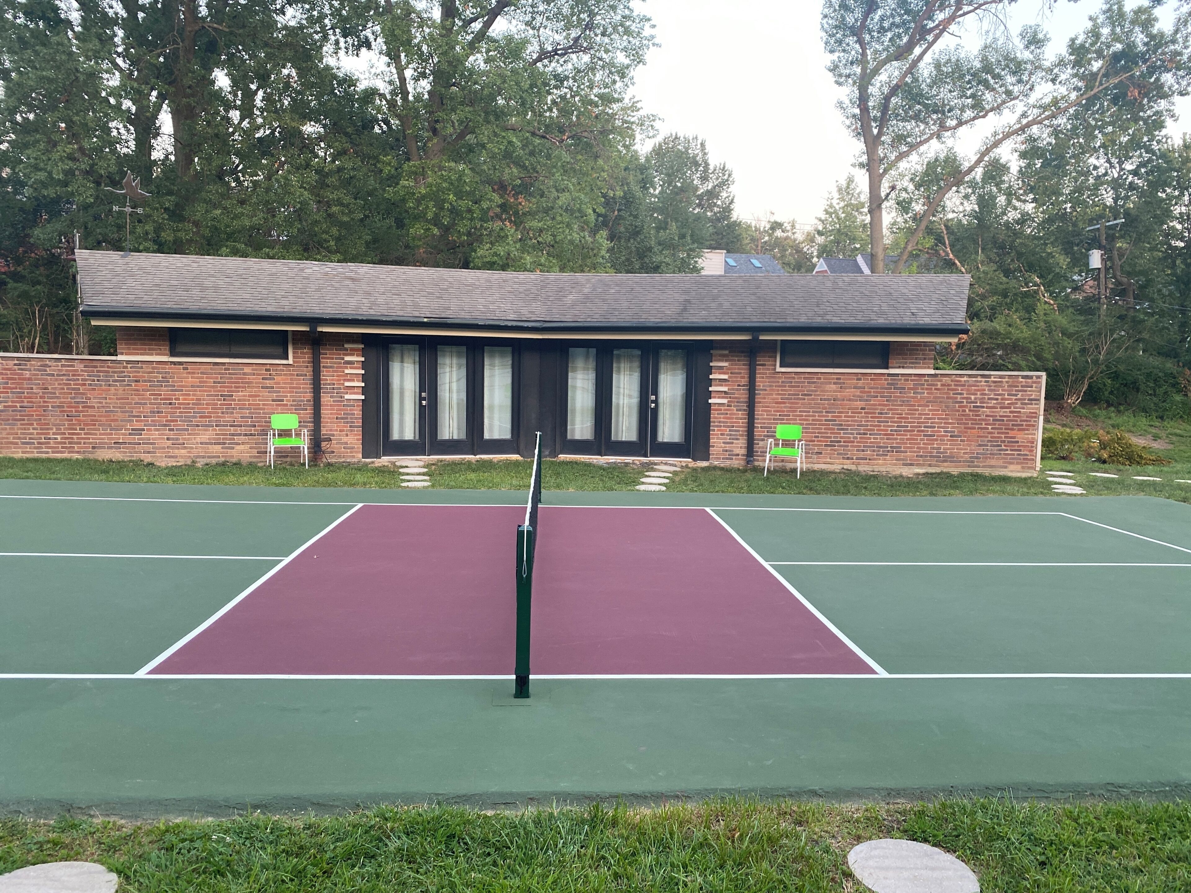 Un court de tennis avec un filet au centre, entouré d’un bâtiment en briques doté de grandes fenêtres et d’une pelouse verdoyante.