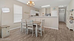 A dining area with a wooden table and chairs, a chandelier, and a kitchen area with cabinets and a refrigerator.