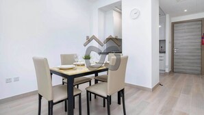 Uma área de refeições com uma mesa de madeira e quatro cadeiras, um relógio na parede e uma porta ao fundo.