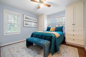 A bedroom with a bed, a bench, a wardrobe, and two windows with blinds.