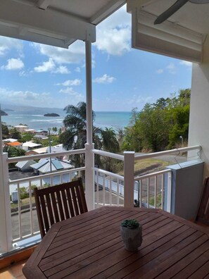 Une table en bois, ornée d’un pot de fleurs, est placée sur un balcon qui offre une vue sur la côte, avec des bâtiments et de la végétation.