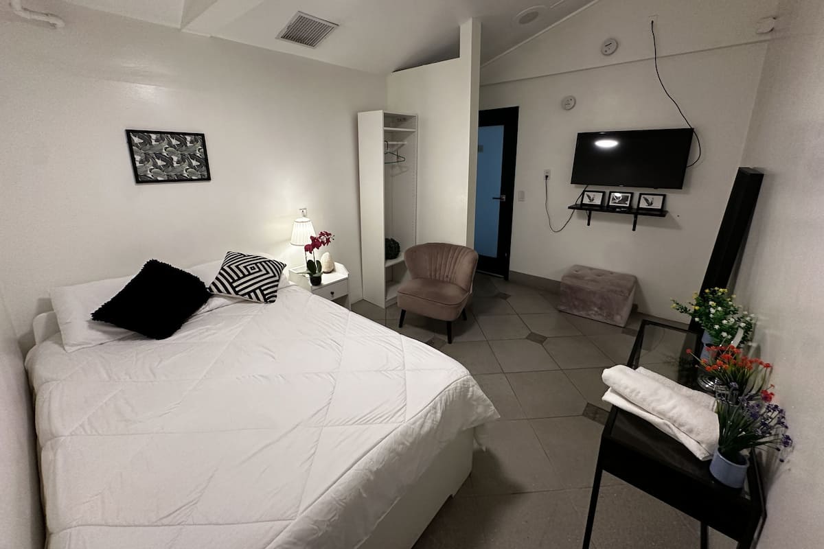 Ein Schlafzimmer mit Bett, Nachttisch, Lampe, Sessel, Fernseher und einem kleinen Tisch mit Blumen.