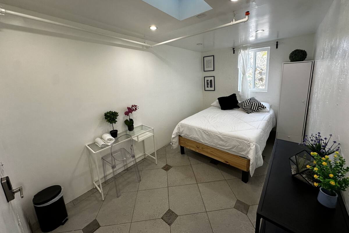 Ein Schlafzimmer mit einem Bett, einem Schreibtisch, einem Stuhl, einem Mülleimer und einem Fenster.