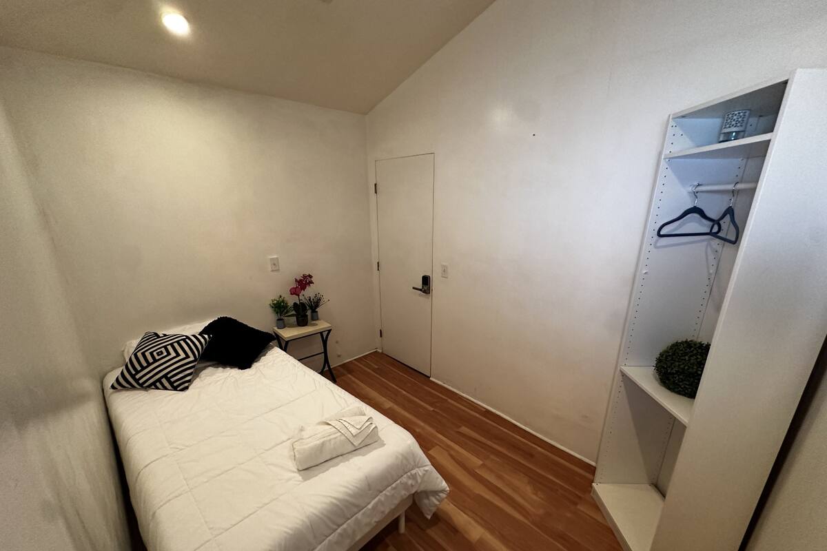 Ein Schlafzimmer mit einem Bett, einem Nachttisch mit Lampe, einer kleinen Pflanze und einem Kleiderschrank mit Kleiderhaken.