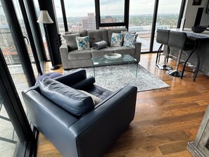 A modern living room with a leather sofa, a grey couch with floral cushions, a glass coffee table, and a view of the cityscape through large windows.