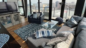 A modern living room with a sofa, a TV, and a large window offering a city view. - 18th floor 2 bed 2 bath overlooking downtown  (Winnipeg)