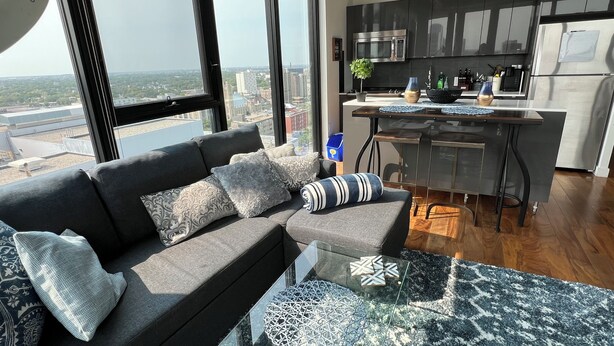 A modern living room with a grey sectional sofa, a glass coffee table, and a view of the city through large windows. - 18th floor 2 bed 2 bath overlooking downtown  (Winnipeg)