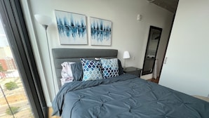 A modern bedroom with a large bed, two abstract paintings, a nightstand, and a window with a view of the city. - 18th floor 2 bed 2 bath overlooking downtown  (Winnipeg)