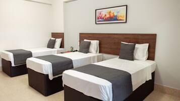 A hotel room with three single beds, each with a wooden headboard, white bedding, and gray throw pillows. A colorful abstract painting hangs on the wall above the beds.