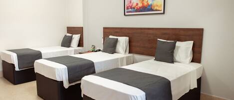 Habitación de hotel con tres camas individuales, cada una con cabecera de madera, ropa de cama blanca y almohadones grises. Un cuadro abstracto y colorido cuelga de la pared sobre las camas.