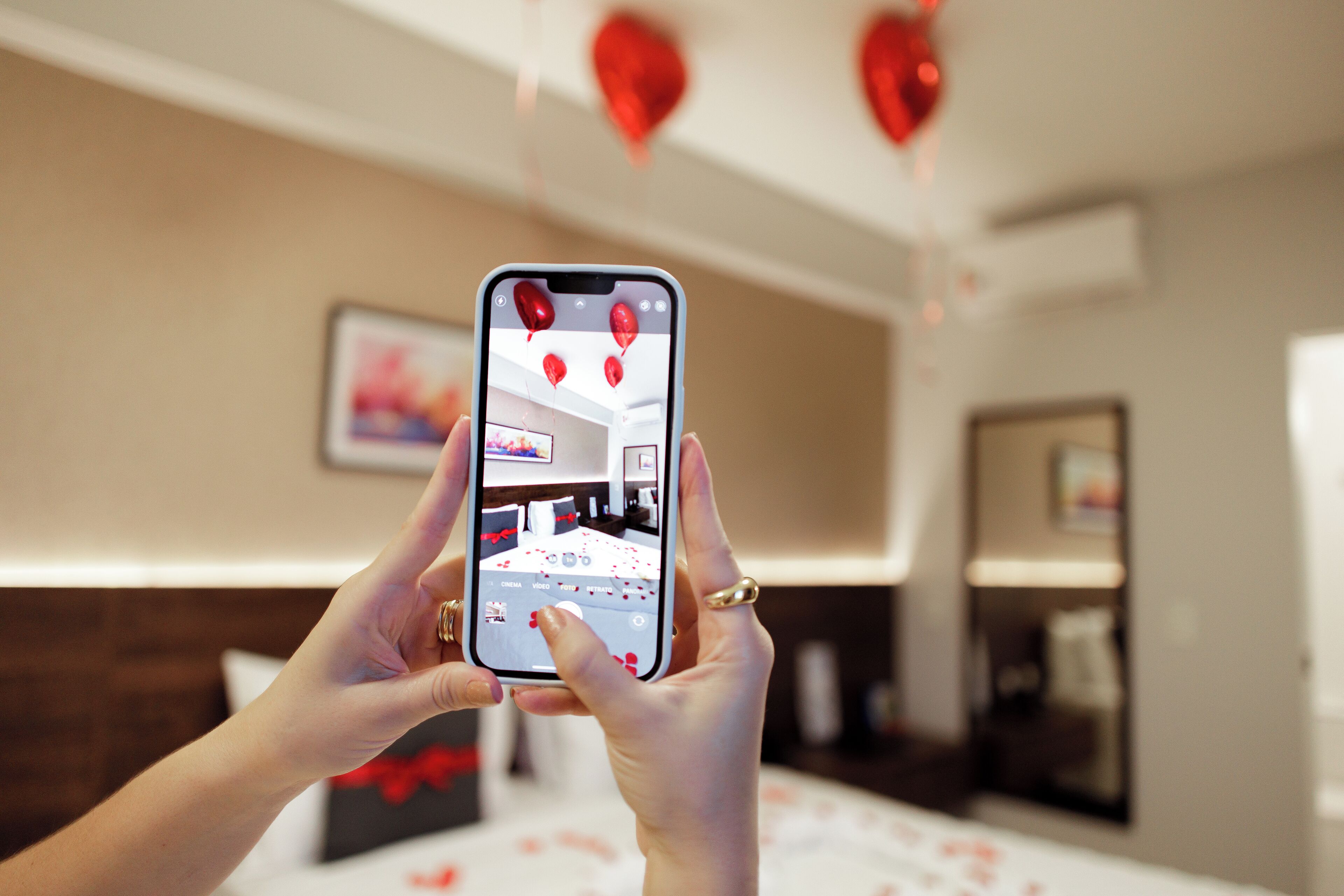 A person holding a smartphone, displaying a room with a bed, a bedside table, and a wall-mounted TV.