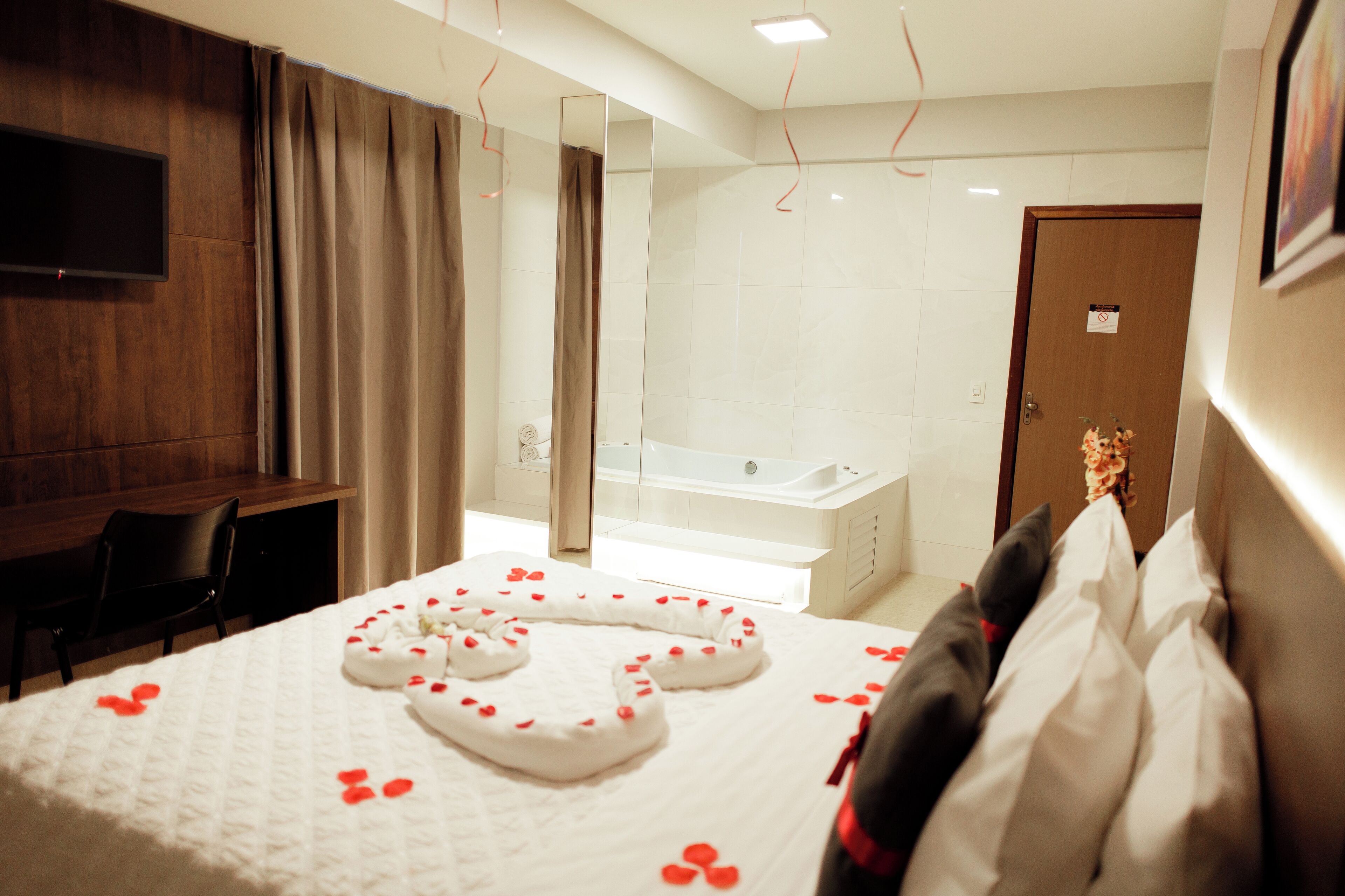 Quarto de hotel com cama, escrivaninha, cadeira, televisão, banheiro com banheira e porta com placa.