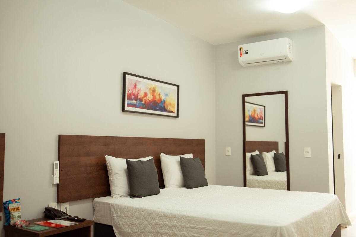 A hotel room with a bed, a bedside table with a phone and a bag, a wall-mounted air conditioner, and a painting.