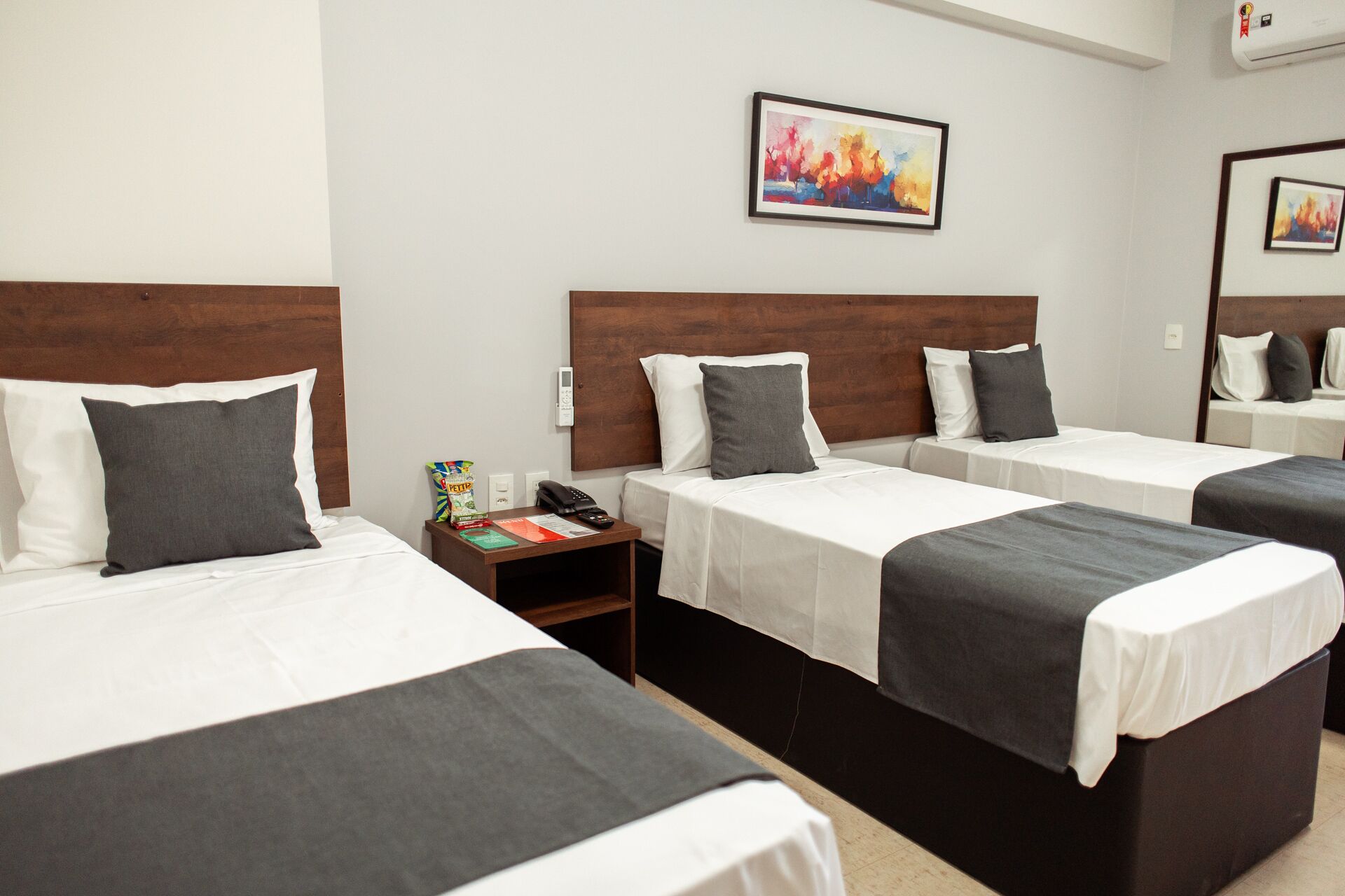 Habitación de hotel con tres camas individuales, cada una con cubrecama blanco y gris, cabecero de madera y almohada gris. Una mesita de noche tiene un teléfono, un paquete de bocadillos y una pequeña mesa con un libro y papeles. Un cuadro abstracto y colorido cuelga en la pared sobre las camas.
