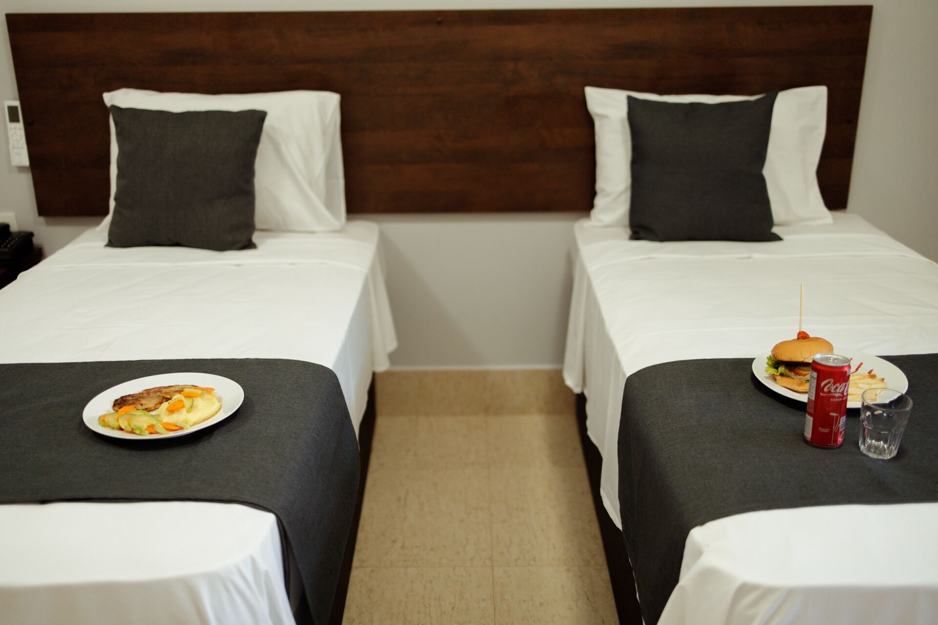 A hotel room with two beds, each with a white bedspread and a dark headboard. There is a plate of food on one bed and a burger with a drink on the other.