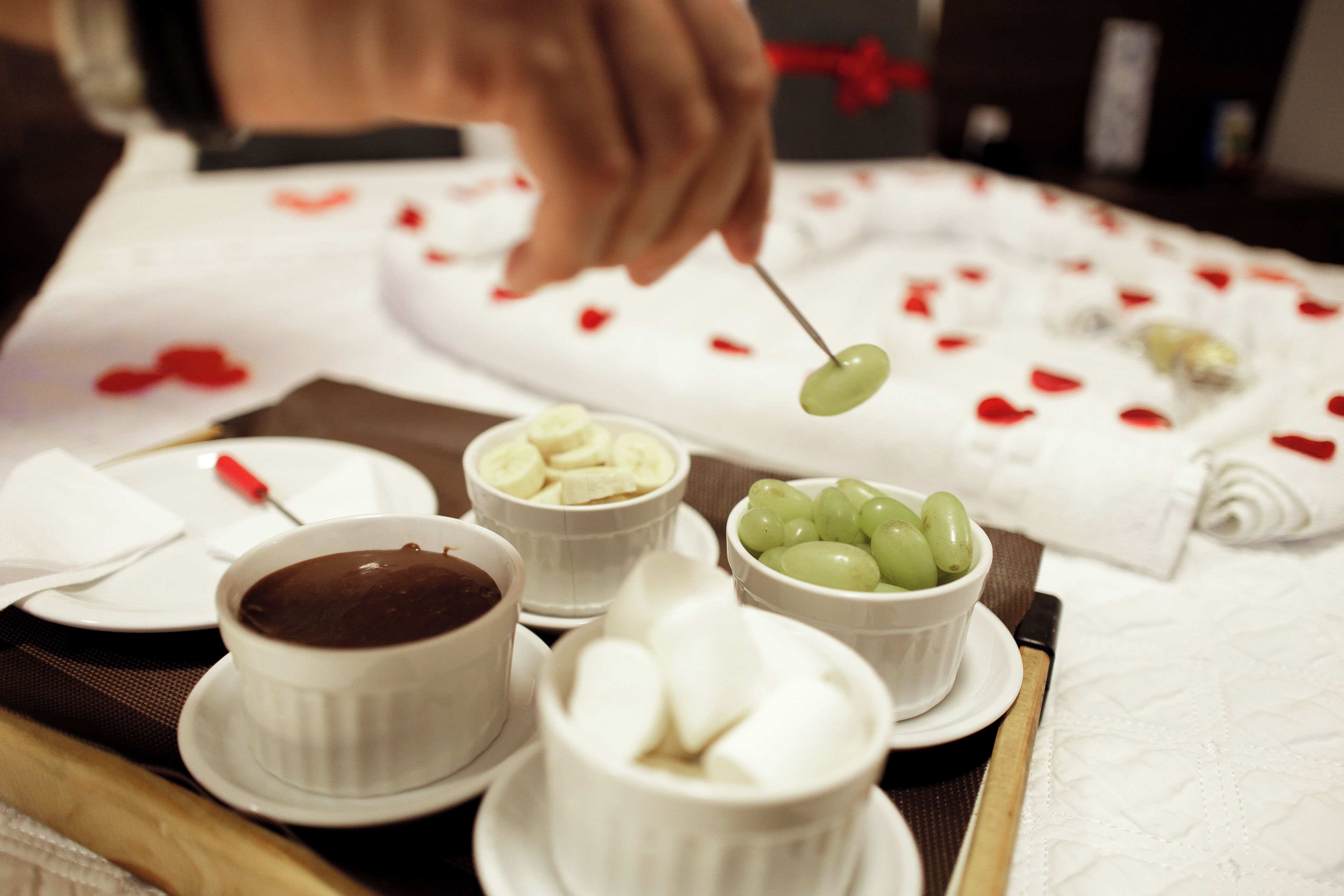 Une table sur laquelle on trouve divers aliments, notamment des bols de chocolat, des bananes et des raisins, et une personne qui trempe un aliment vert dans le chocolat.