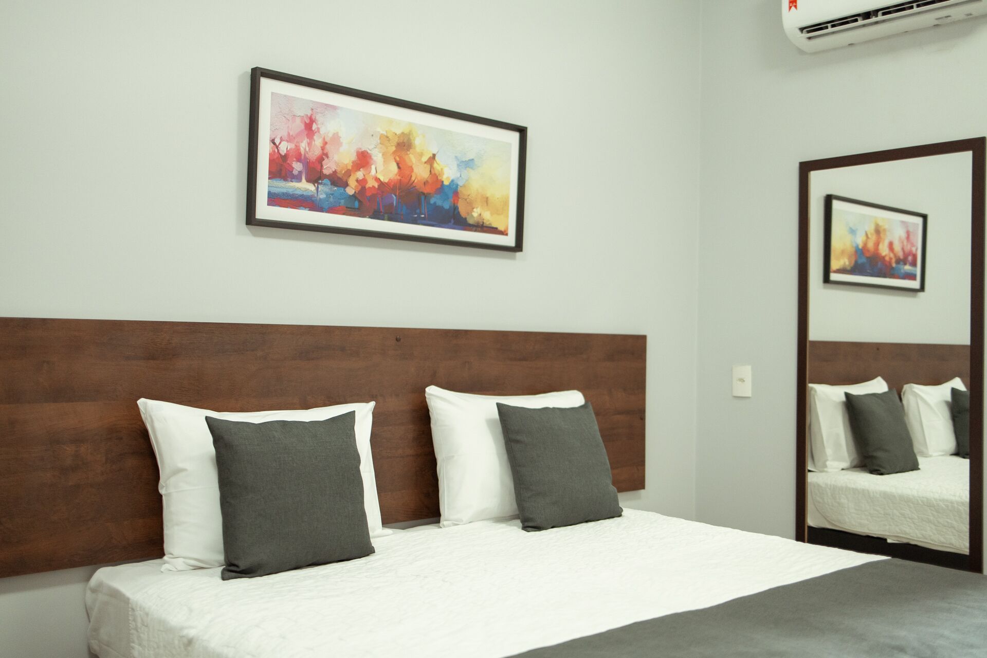 A hotel room with a wooden headboard, a bed with white and gray bedding, two pillows, and a framed abstract painting above the bed.
