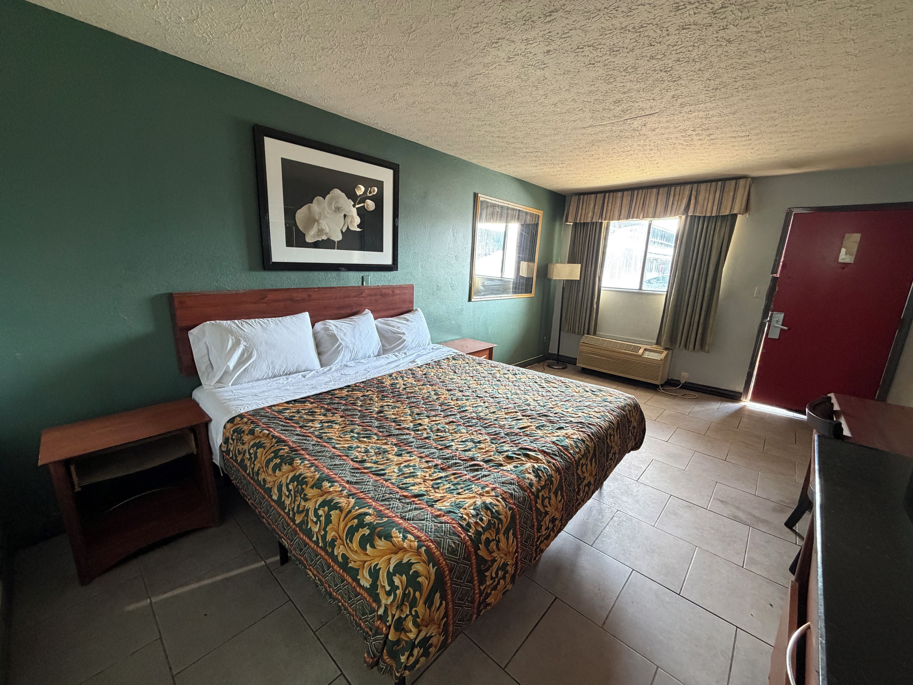 A hotel room with a large bed, a nightstand, a desk, a chair, a framed picture on the wall, and a window with curtains.