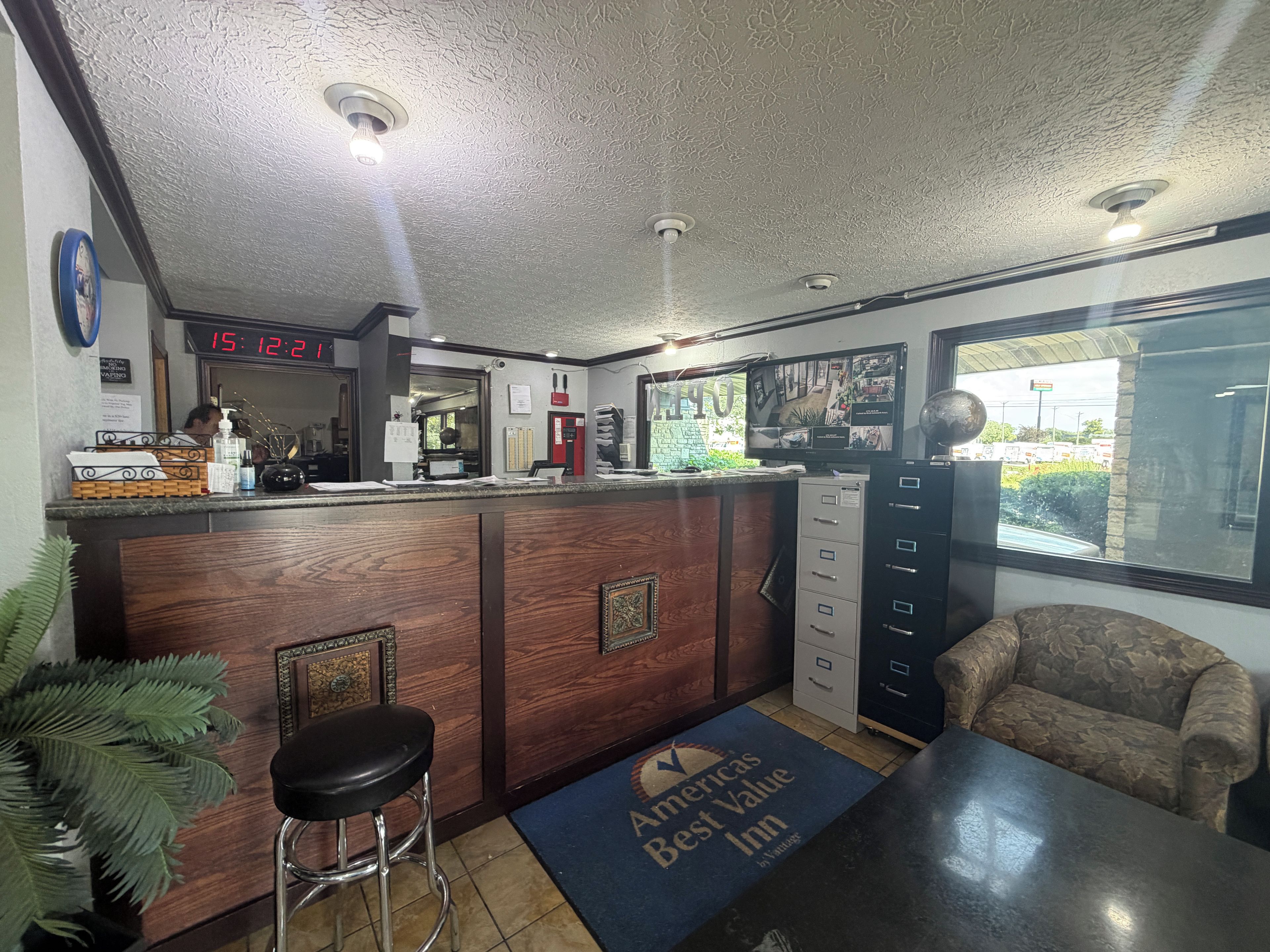 A reception area with a wooden counter, a digital clock, a desk with a computer, a chair, and a view of the outside through a window.