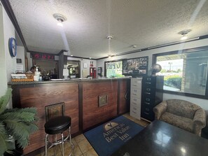 A reception area with a wooden counter, a digital clock, a desk with a computer, a chair, and a view of the outside through a window.