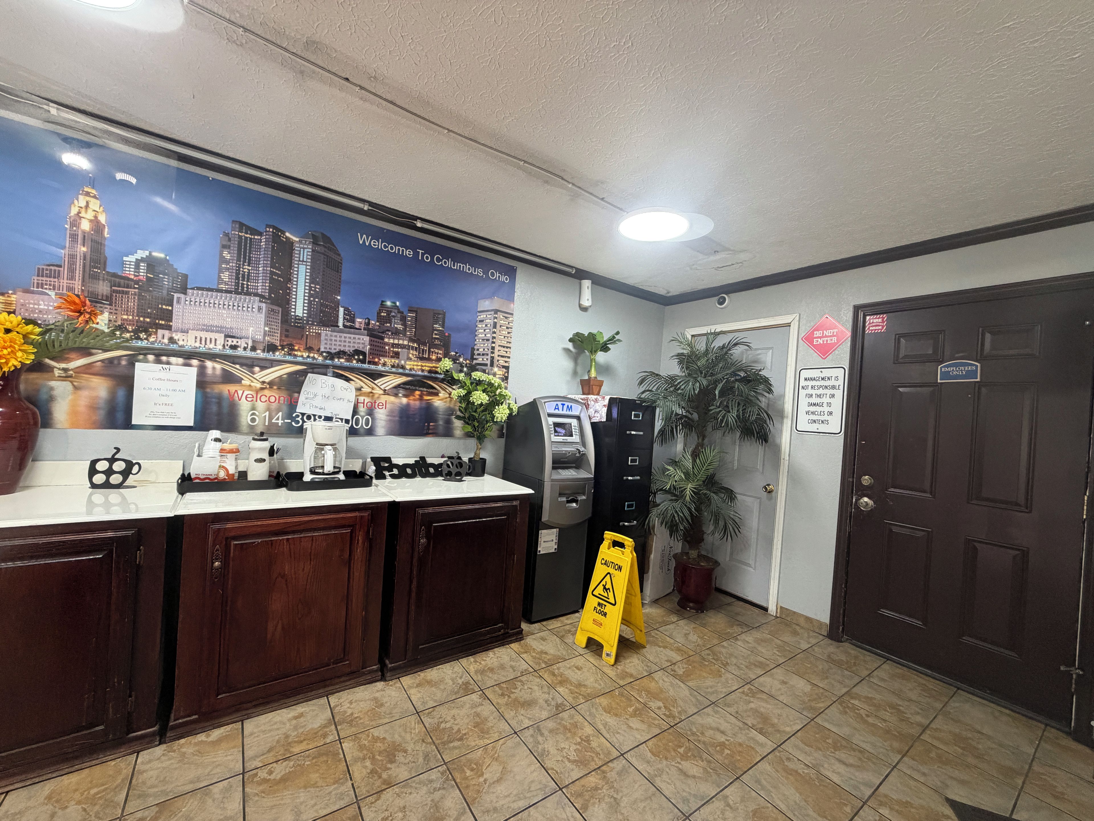 A hotel reception area with a cityscape poster, an ATM, a coffee station, and a caution wet floor sign.