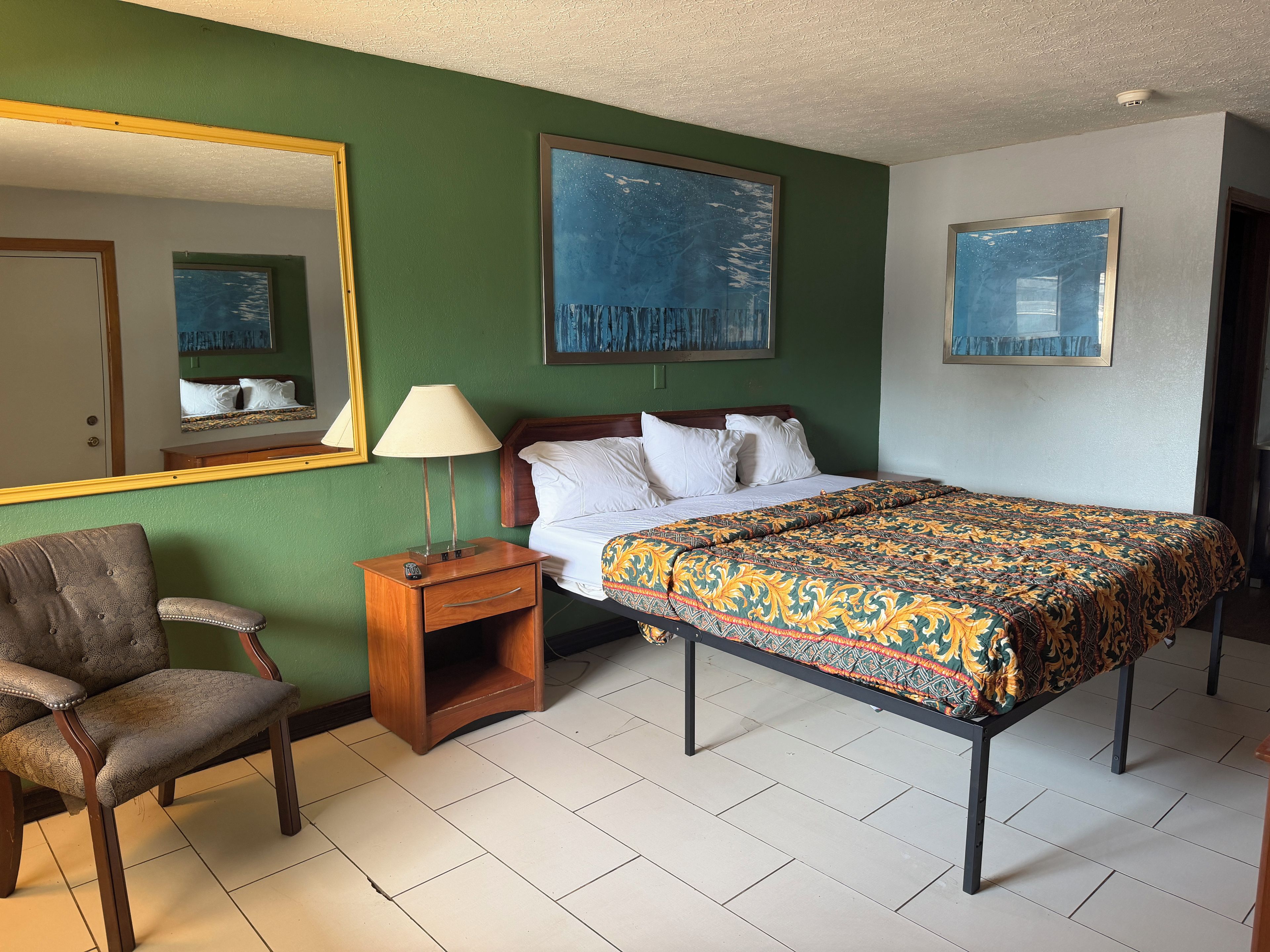 A hotel room with a bed, a chair, a nightstand, a lamp, and two paintings on the wall.