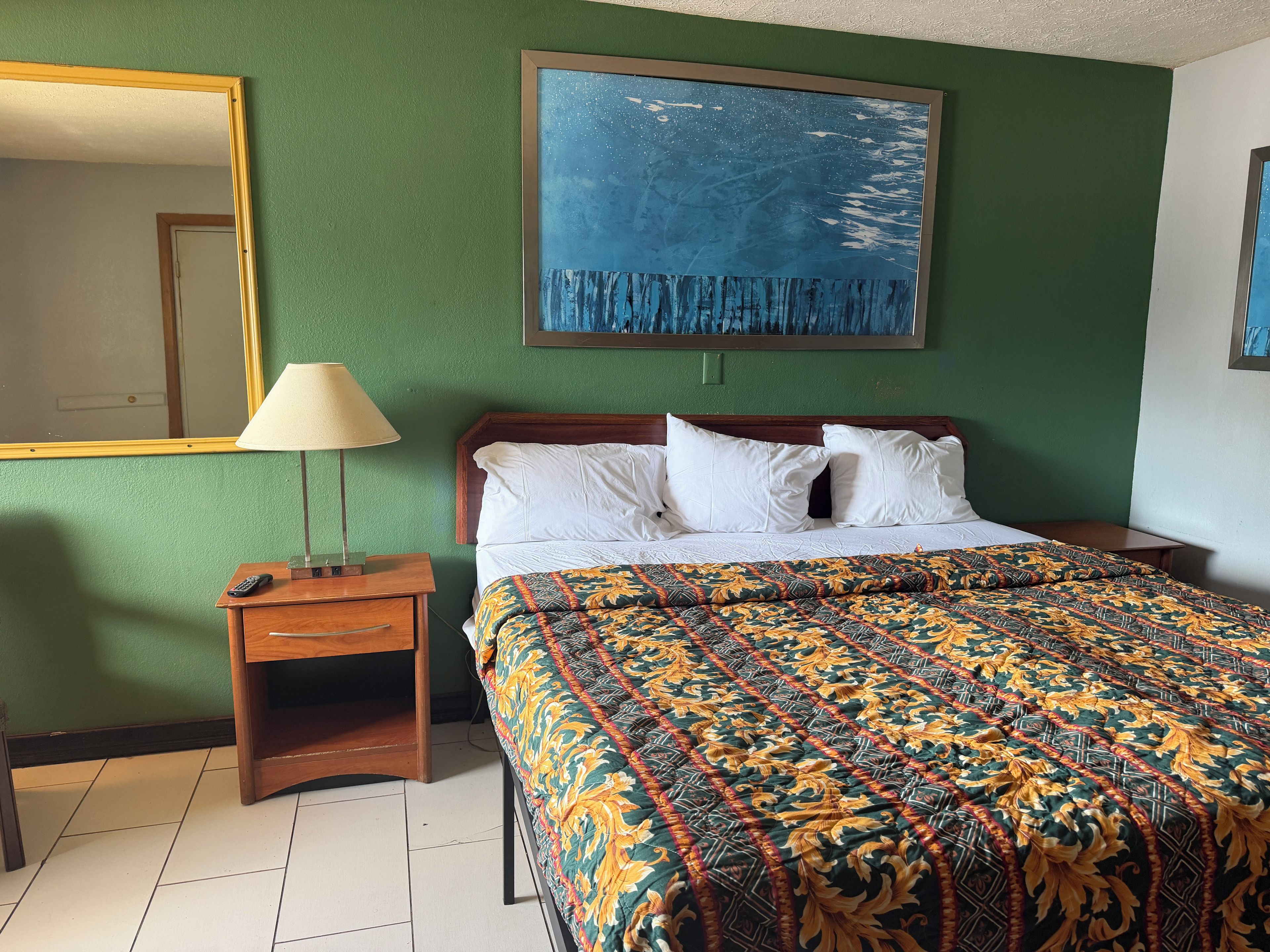 A hotel room with a bed, a nightstand, a lamp, a mirror, and a painting on the wall.
