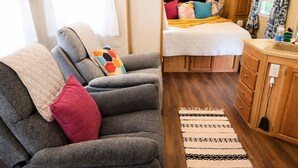 Un interior de autocaravana acogedor, con un sofá gris, una cama con almohadones de colores, armarios de madera y una alfombra estampada.