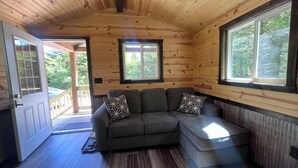 A cozy cabin interior with a wooden ceiling, a gray sectional sofa with patterned cushions, and large windows offering a view of the outdoors. - Quiet Cabin in the woods.  (Williamsburg)