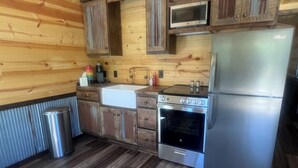 A compact kitchen with wooden walls, a stainless steel refrigerator, a microwave, a stove, and a sink. - Quiet Cabin in the woods.  (Williamsburg)