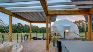 Una terraza de madera con jacuzzi, mobiliario exterior y vistas a una estructura geodésica.
