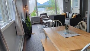 En moderne stue med et træspisebord, hvide stole, en sofa og udsigt til bjerge gennem store vinduer.