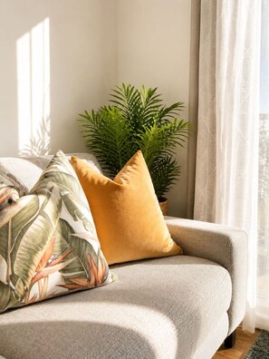 Un canapé avec un seul coussin jaune et un oreiller à motifs, placé à côté d’une plante verte dans une pièce avec une fenêtre.