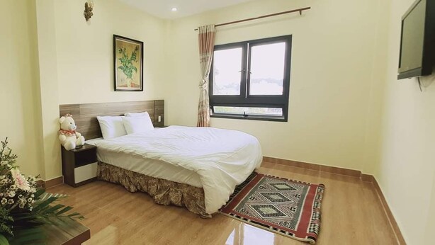 A bedroom with a bed, a nightstand, a teddy bear, a window, and a TV on the wall. - Ngoc Tran Hostel (Cam Ly - Da Lat)