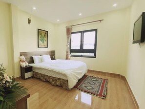 A bedroom with a bed, a nightstand, a teddy bear, a window, and a TV on the wall. - Ngoc Tran Hostel (Da Lat)