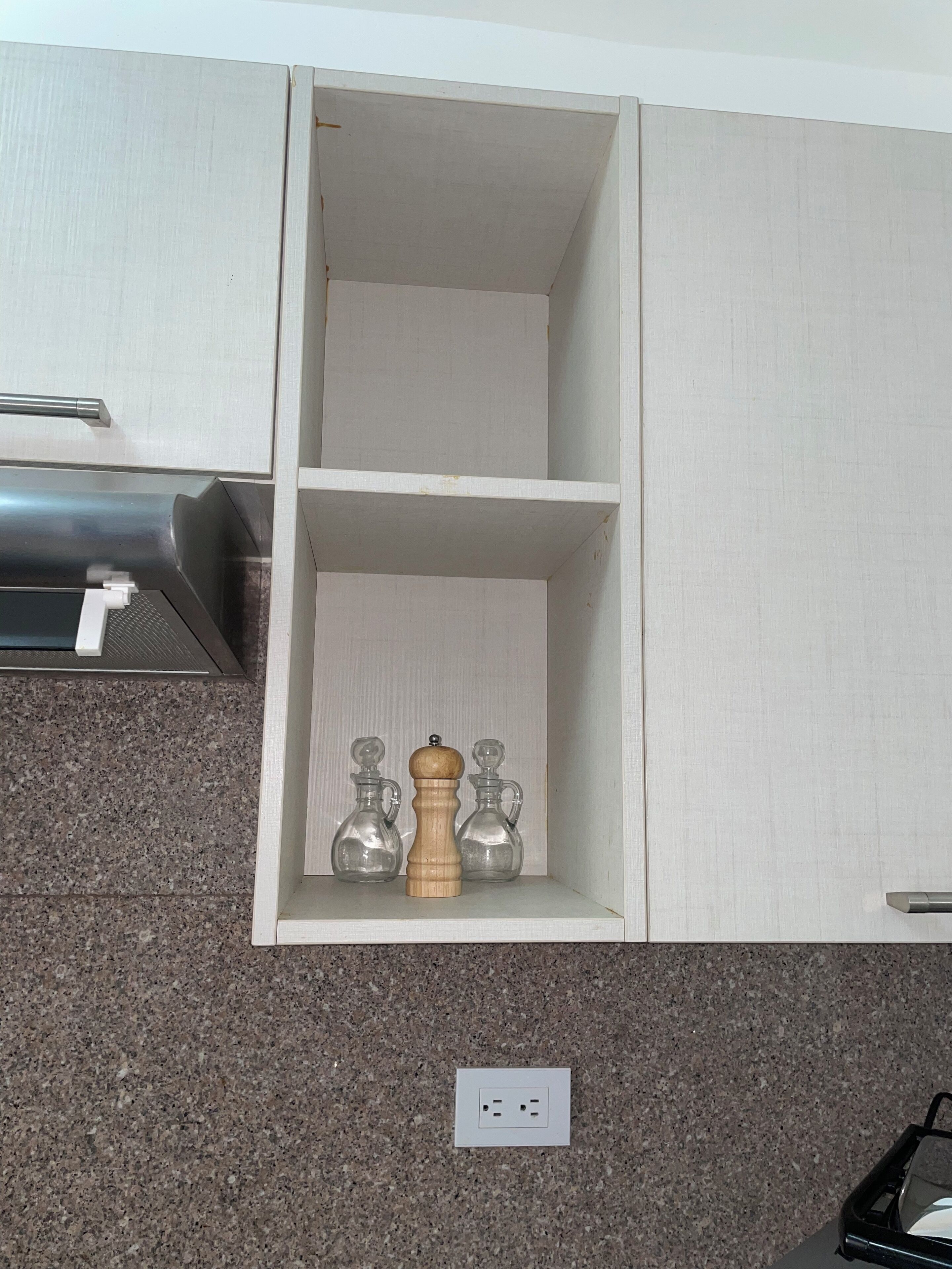 Una cocina con encimera de granito, estufa integrada y un armario blanco con un estante que contiene dos saleros y pimenteros de vidrio, además de un molinillo de pimienta de madera.