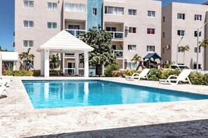 A swimming pool with lounge chairs, a covered pavilion, and a multi-story building with balconies. - Hermoso Apartamento con Piscina (La Romana)