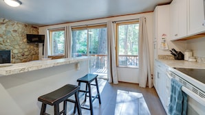 A kitchen with a stone wall, a TV, a bar counter, and a view of trees through large windows. - Peaceful Leavenworth Cabin with Deck & Fire Pit! (Leavenworth)