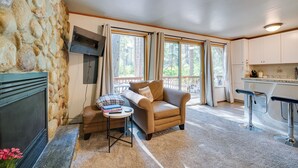 A cozy living room with a stone wall, a fireplace, a sofa, and a kitchen area with a bar. - Peaceful Leavenworth Cabin with Deck & Fire Pit! (Leavenworth)