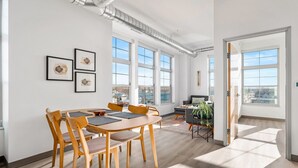 A spacious room with a large dining table, multiple chairs, and a sofa. There are three large windows providing natural light and a view of the city. - Oak Tree House 305 (Traverse City)