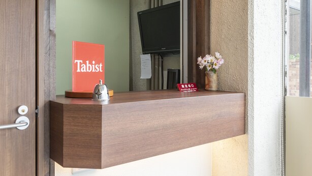 Una recepción de hotel con un mostrador de madera, un letrero 'Tabist', un timbre, un jarrón con flores y un televisor de pantalla plana al fondo.