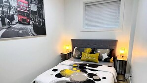 A bedroom with a bed, a headboard, bedside tables, a wall-mounted lamp, a framed picture, and a window with blinds.