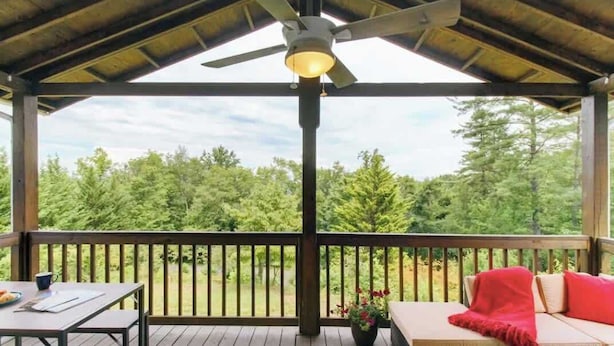 Un espace extérieur abrité, équipé d’une table et de sièges, d’un ventilateur de plafond et offrant une vue sur les arbres.